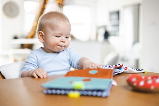 Happy infant sitting at dining table and playing with his toy in traditional scandinavian designer wooden high chair in modern bright atic home. Cute baby playing with toys.