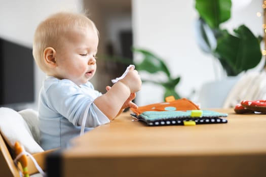 Happy infant sitting at dining table and playing with his toy in traditional scandinavian designer wooden high chair in modern bright atic home. Cute baby playing with toys.