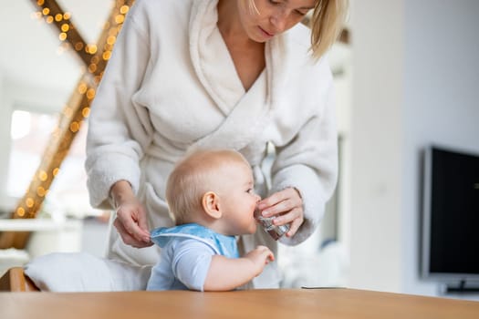 Casual domestic lifestyle moments concept. Infant baby boy drinking water from small glass with a help of his mother wearing cosy bathrope