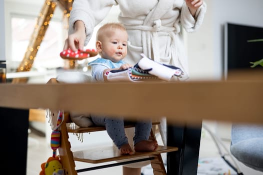 Happy infant sitting at dining table and playing with his toy in traditional scandinavian designer wooden high chair in modern bright atic home superwised by his mother.