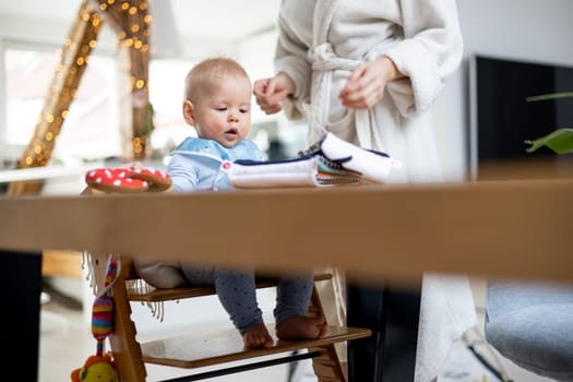 Happy infant sitting at dining table and playing with his toy in traditional scandinavian designer wooden high chair in modern bright atic home superwised by his mother.