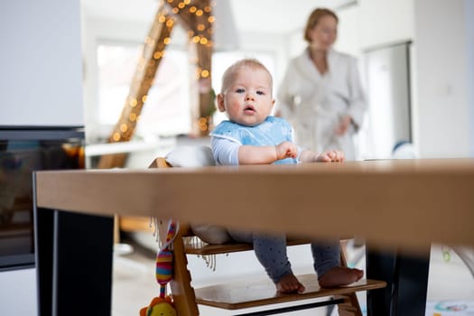 Happy infant sitting at dining table and playing with his toy in traditional scandinavian designer wooden high chair in modern bright atic home superwised by his mother.