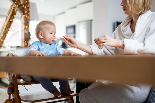 Mother wearing cosy bathrope spoon feeding her baby boy child in baby high chair with fruit puree at dinning table at home. Baby solid food introduction concept.