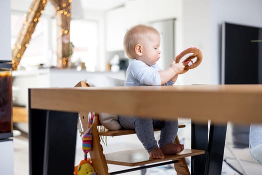 Happy infant sitting at dining table and playing with his toy in traditional scandinavian designer wooden high chair in modern bright atic home. Cute baby playing with toys.