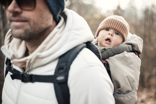 Sporty father carrying his infant son wearing winter jumpsuit and cap in backpack carrier hiking in autumn forest