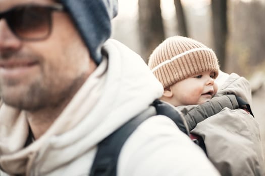 Sporty father carrying his infant son wearing winter jumpsuit and cap in backpack carrier hiking in autumn forest