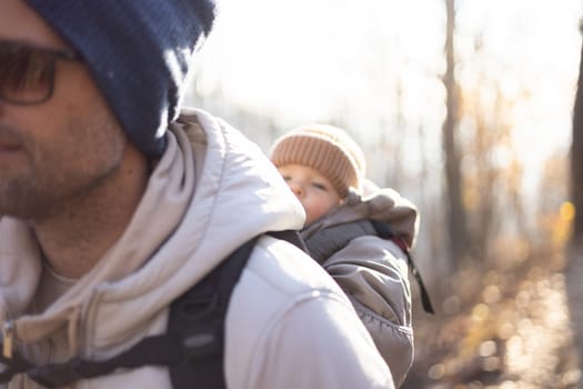 Sporty father carrying his infant son wearing winter jumpsuit and cap in backpack carrier hiking in autumn forest