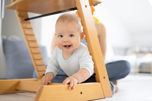 Happy infant climbing under traditional scandinavian designer wooden high chair and in modern bright home. Cute baby smiling in camera.
