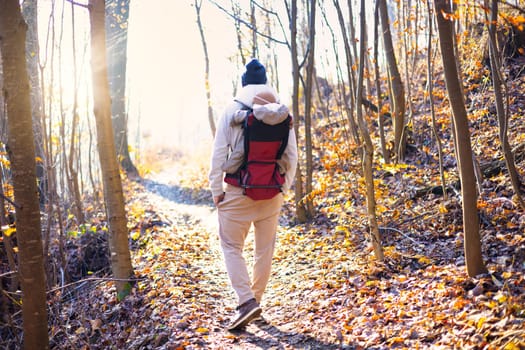 Sporty father carrying his infant son wearing winter jumpsuit and cap in backpack carrier hiking in autumn forest