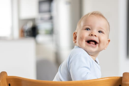 Happy infant sitting in traditional scandinavian designer wooden high chair and laughing out loud in modern bright home. Cute baby smile