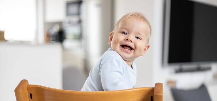 Happy infant sitting in traditional scandinavian designer wooden high chair and laughing out loud in modern bright home. Cute baby smile