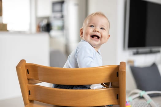 Happy infant sitting in traditional scandinavian designer wooden high chair and laughing out loud in modern bright home. Cute baby smile
