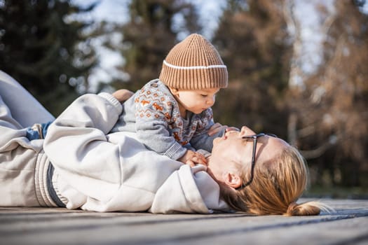 Happy family. Young mother playing with her baby boy infant oudoors on sunny autumn day. Portrait of mom and little son on wooden platform by lake. Positive human emotions, feelings, joy