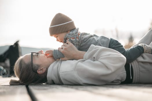 Happy family. Young mother playing with her baby boy infant oudoors on sunny autumn day. Portrait of mom and little son on wooden platform by lake. Positive human emotions, feelings, joy