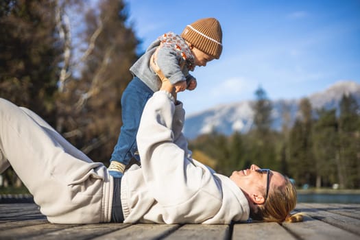 Happy family. Young mother playing with her baby boy infant oudoors on sunny autumn day. Portrait of mom and little son on wooden platform by lake. Positive human emotions, feelings, joy