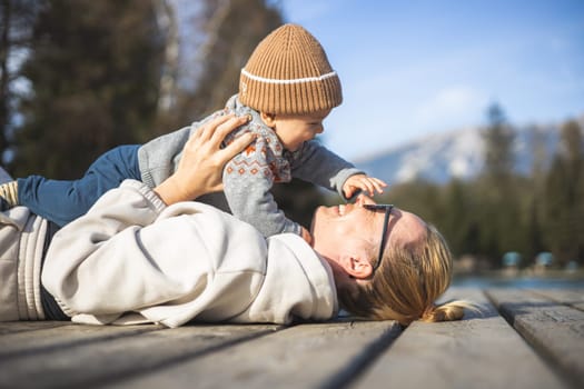 Happy family. Young mother playing with her baby boy infant oudoors on sunny autumn day. Portrait of mom and little son on wooden platform by lake. Positive human emotions, feelings, joy