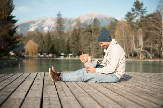 Happy family. Father playing with her baby boy infant oudoors on sunny autumn day. Portrait of dad and little son on wooden platform by lake. Positive human emotions, feelings, joy