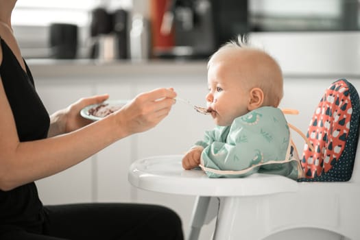 Mother spoon feeding her baby boy child in baby chair with fruit puree in kitchen at home. Baby solid food introduction concept