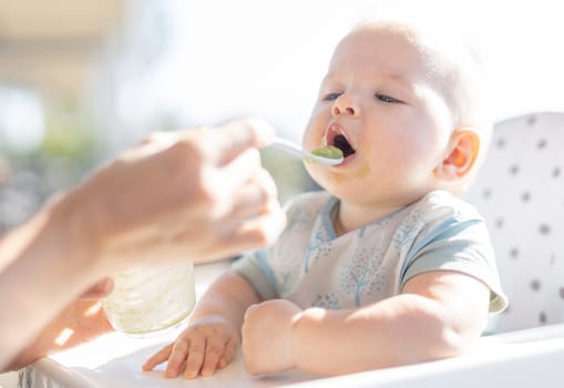 Mother spoon feeding her baby boy child in baby chair with fruit puree on a porch on summer vacations. Baby solid food introduction concept