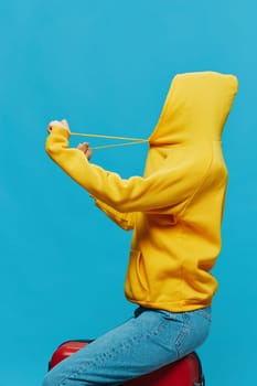 Smile woman sitting on a suitcase in a yellow hoodie, blue jeans and glasses on a blue background, packing for a trip. High quality photo