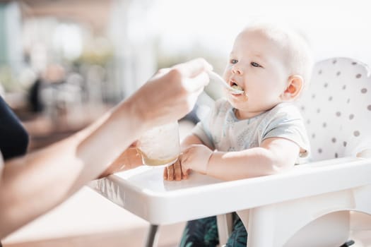 Mother spoon feeding her baby boy child in baby chair with fruit puree on a porch on summer vacations. Baby solid food introduction concept
