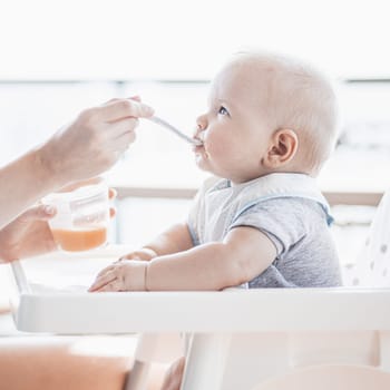 Mother spoon feeding her baby boy child in baby chair with fruit puree on a porch on summer vacations. Baby solid food introduction concept