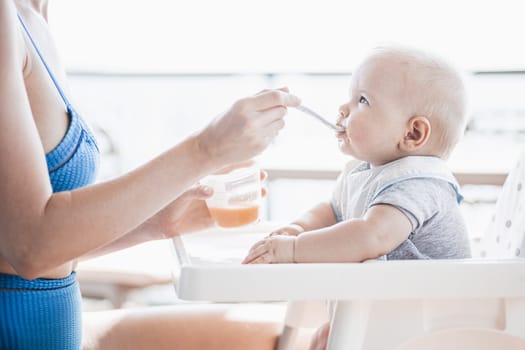 Mother spoon feeding her baby boy child in baby chair with fruit puree on a porch on summer vacations. Baby solid food introduction concept