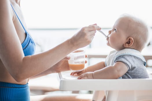 Mother spoon feeding her baby boy child in baby chair with fruit puree on a porch on summer vacations. Baby solid food introduction concept