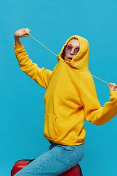 Smile woman sitting on a suitcase in a yellow hoodie, blue jeans and glasses on a blue background, packing for a trip. High quality photo