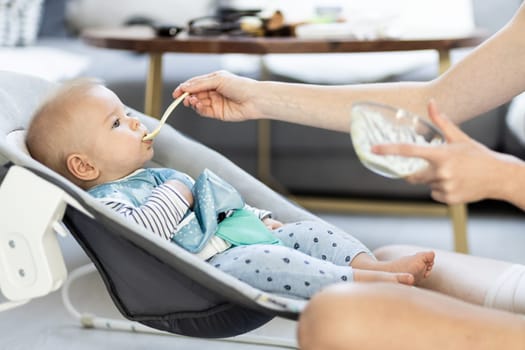 Mother spoon feeding her baby boy child in baby chair with fruit puree. Baby solid food introduction concept