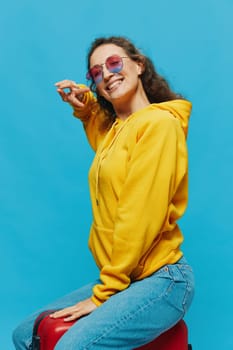 Smile woman sitting on a suitcase in a yellow hoodie, blue jeans and glasses on a blue background, packing for a trip. High quality photo