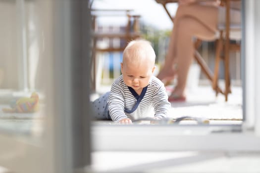Cute little infant baby boy playing with toys outdoors at the patio in summer being supervised by her mother seen in the background. Selective focus.