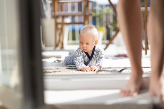 Cute little infant baby boy playing with toys outdoors at the patio in summer being supervised by her mother seen in the background. Selective focus.