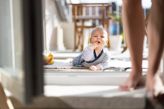 Cute little infant baby boy playing with toys outdoors at the patio in summer being supervised by her mother seen in the background. Selective focus.