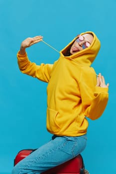 Smile woman sitting on a suitcase in a yellow hoodie, blue jeans and glasses on a blue background, packing for a trip. High quality photo