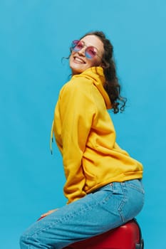 Smile woman sitting on a suitcase in a yellow hoodie, blue jeans and glasses on a blue background, packing for a trip. High quality photo