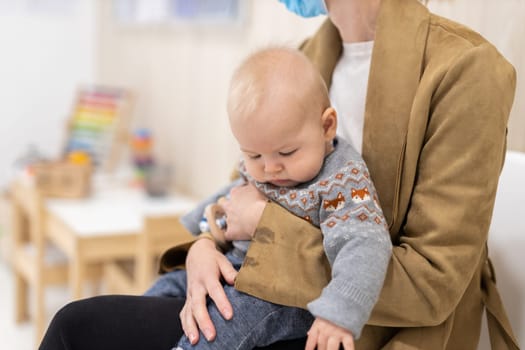 Mother holding infant baby boy in her lap, sitting and waiting in front of doctor's office for pediatric well check. child's health care concept.