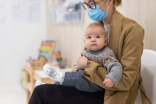 Mother holding infant baby boy in her lap, sitting and waiting in front of doctor's office for pediatric well check. child's health care concept.