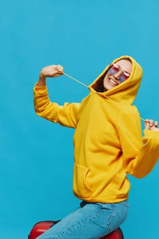 Smile woman sitting on a suitcase in a yellow hoodie, blue jeans and glasses on a blue background, packing for a trip. High quality photo