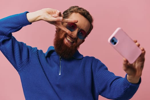 Portrait of a redheaded man with phone in hand taking selfies and photos on his phone with a smile on a pink background, blogger. High quality photo