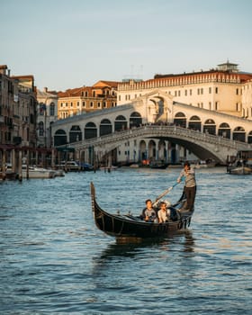 Beautiful view of tourists sitting in gondola with a gondolier, floating along canal on the backdrop of Rialto bridge on sunny warm summer evening. Concept of tourism in the romantic city of Italy.