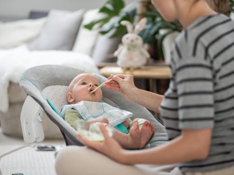 Mother spoon feeding her baby boy child in baby chair with fruit puree. Baby solid food introduction concept