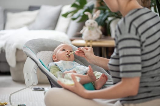 Mother spoon feeding her baby boy child in baby chair with fruit puree. Baby solid food introduction concept