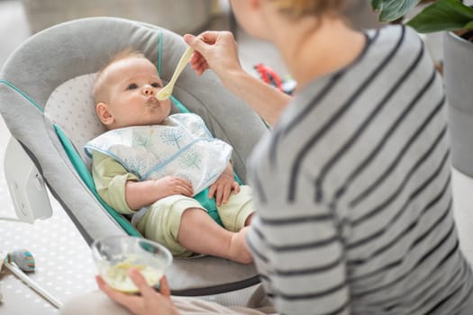 Mother spoon feeding her baby boy child in baby chair with fruit puree. Baby solid food introduction concept