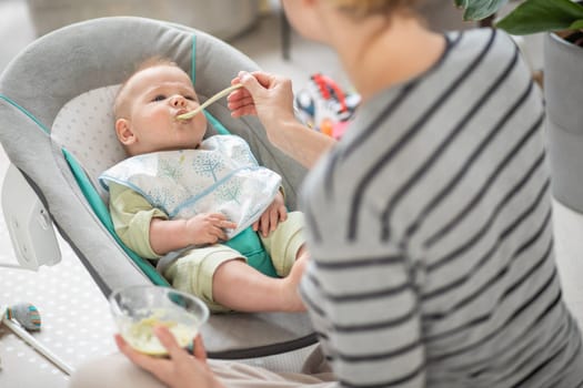 Mother spoon feeding her baby boy child in baby chair with fruit puree. Baby solid food introduction concept