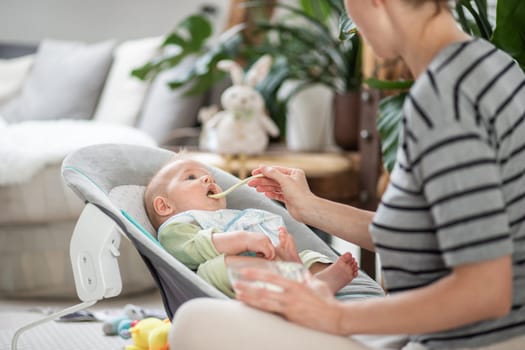 Mother spoon feeding her baby boy child in baby chair with fruit puree. Baby solid food introduction concept