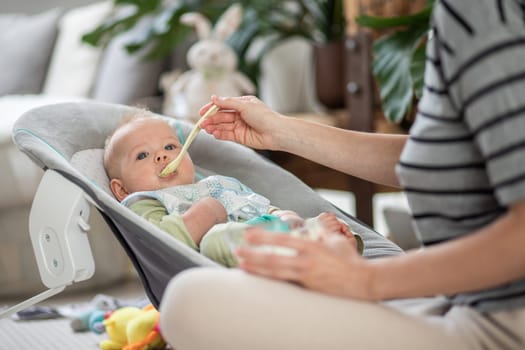 Mother spoon feeding her baby boy child in baby chair with fruit puree. Baby solid food introduction concept