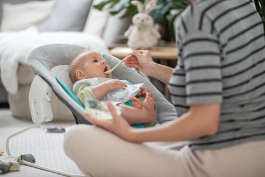 Mother spoon feeding her baby boy child in baby chair with fruit puree. Baby solid food introduction concept