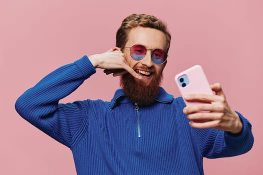 Portrait of a redheaded man with phone in hand taking selfies and photos on his phone with a smile on a pink background, blogger. High quality photo