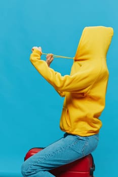 Smile woman sitting on a suitcase in a yellow hoodie, blue jeans and glasses on a blue background, packing for a trip. High quality photo